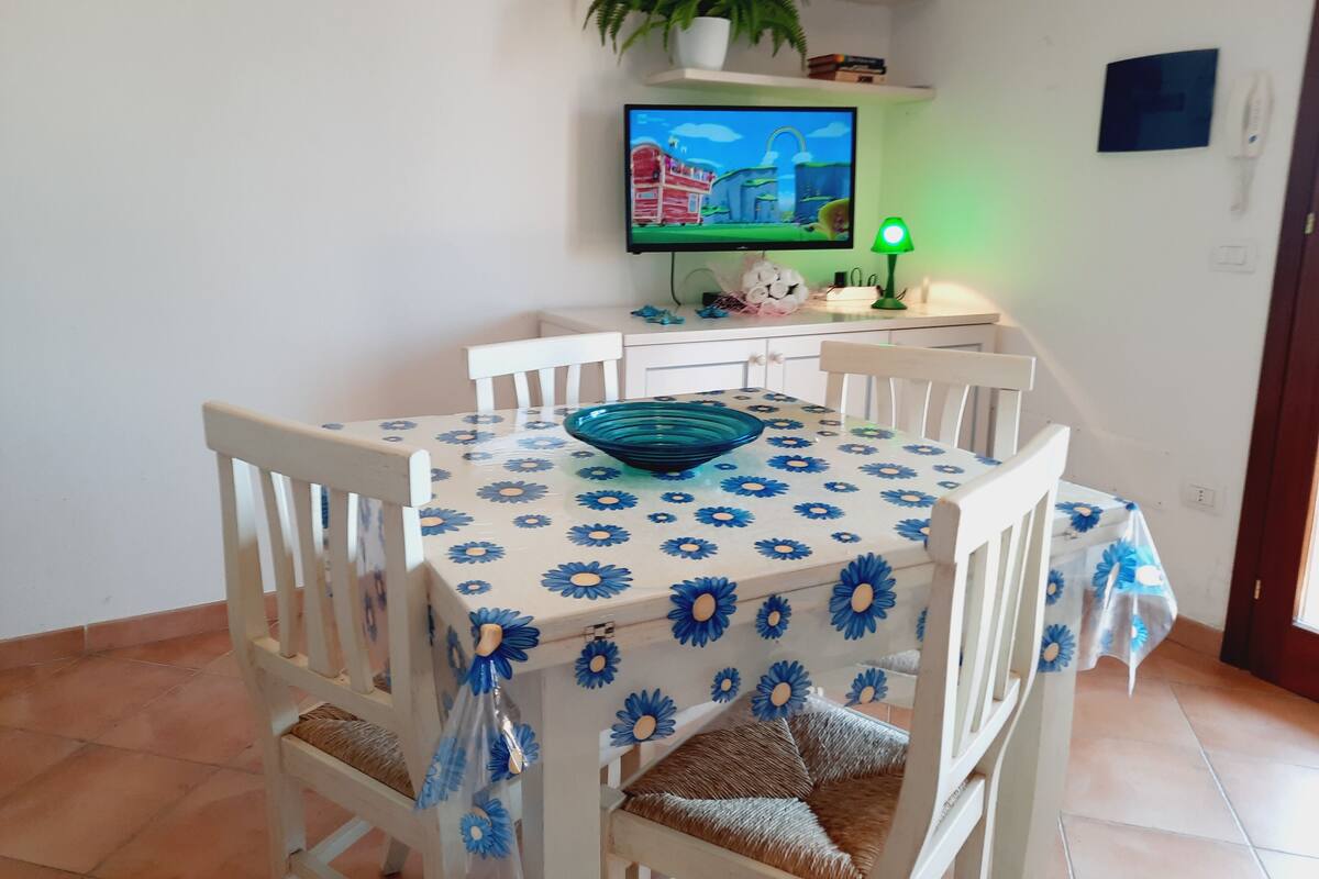 A dining area with a table covered in a blue and white floral tablecloth, surrounded by white chairs with woven seat cushions. A green desk lamp is on the table, and a television is mounted on the wall above it.