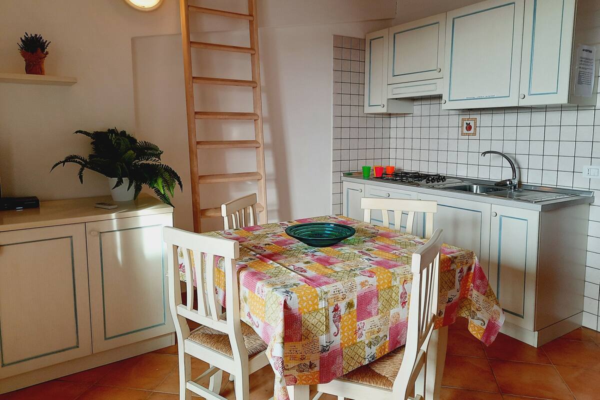 A compact kitchen with white cabinets, a tiled backsplash, a wooden ladder, and a table with a colorful quilt.