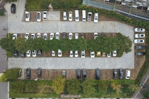 A parking lot with multiple rows of parked cars, surrounded by trees and a building in the background.