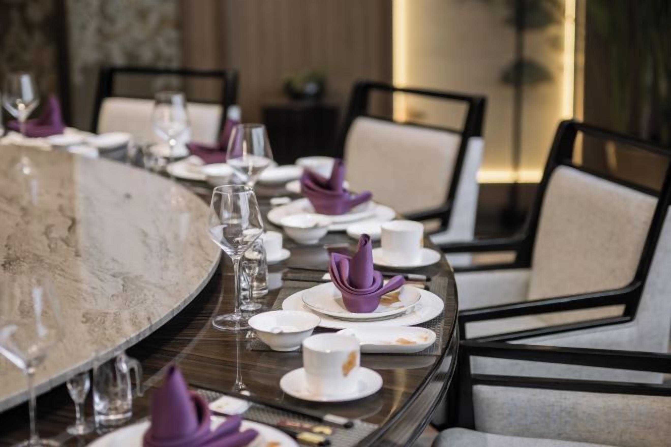 Een eettafel, gedekt met witte borden, paarse servetten en glazen, omringd door stoelen.