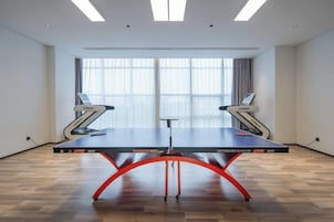 Una mesa de ping pong en una habitación con ventanas amplias, una cinta de correr y un escritorio.