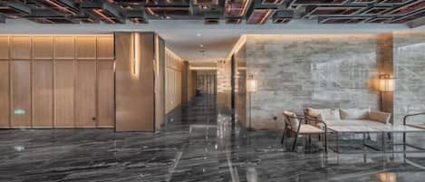 A modern hotel lobby with a reflective marble floor, a seating area with chairs and a table, and a wooden partition wall.