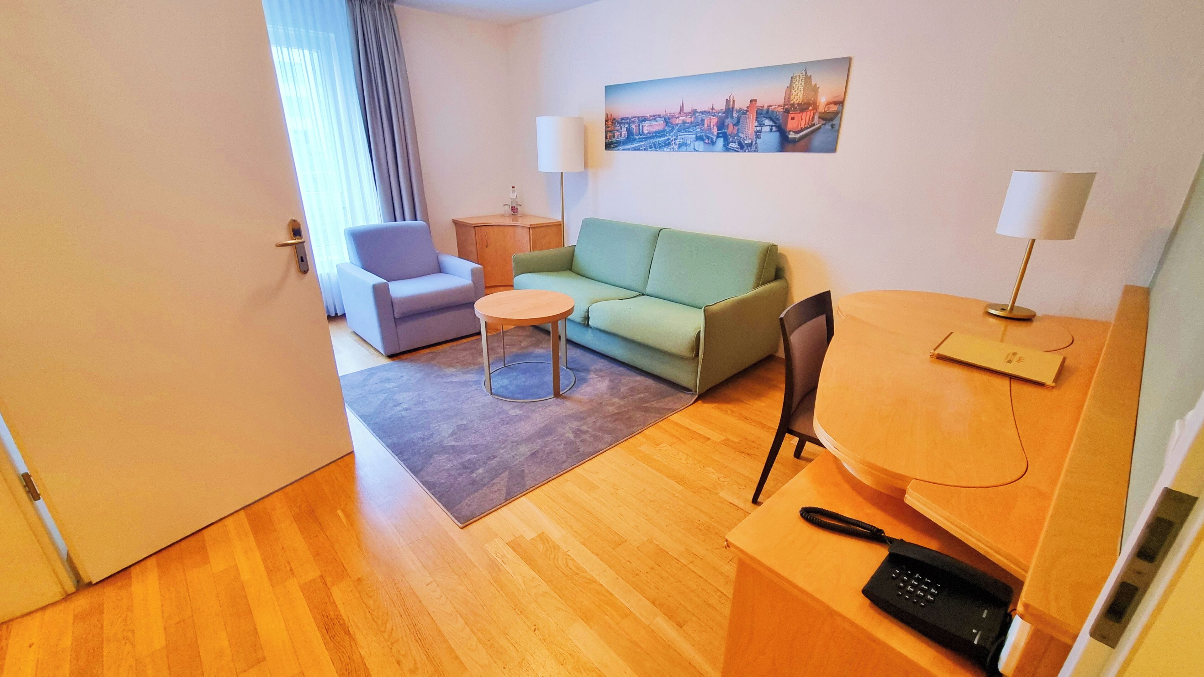 A hotel room with a green sofa, a small round table, a chair, a desk with a lamp, and a painting of a cityscape on the wall.