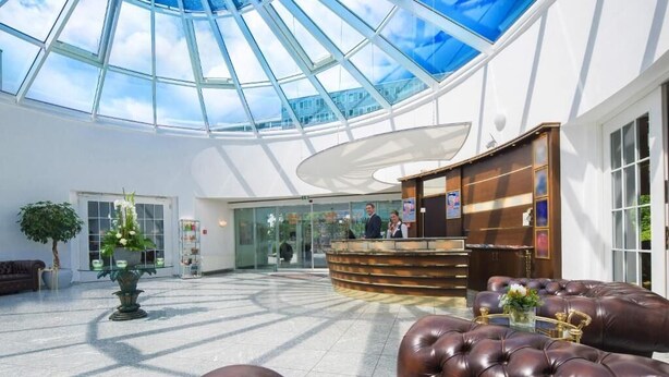 A spacious lobby with a glass ceiling, a reception desk, leather seating, and a potted plant. - New Living Home Residenzhotel Hamburg (Hamburg)