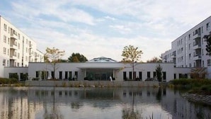 A modern building with a central glass facade, surrounded by a calm water body and greenery. - New Living Home Residenzhotel Hamburg (Hamburg)