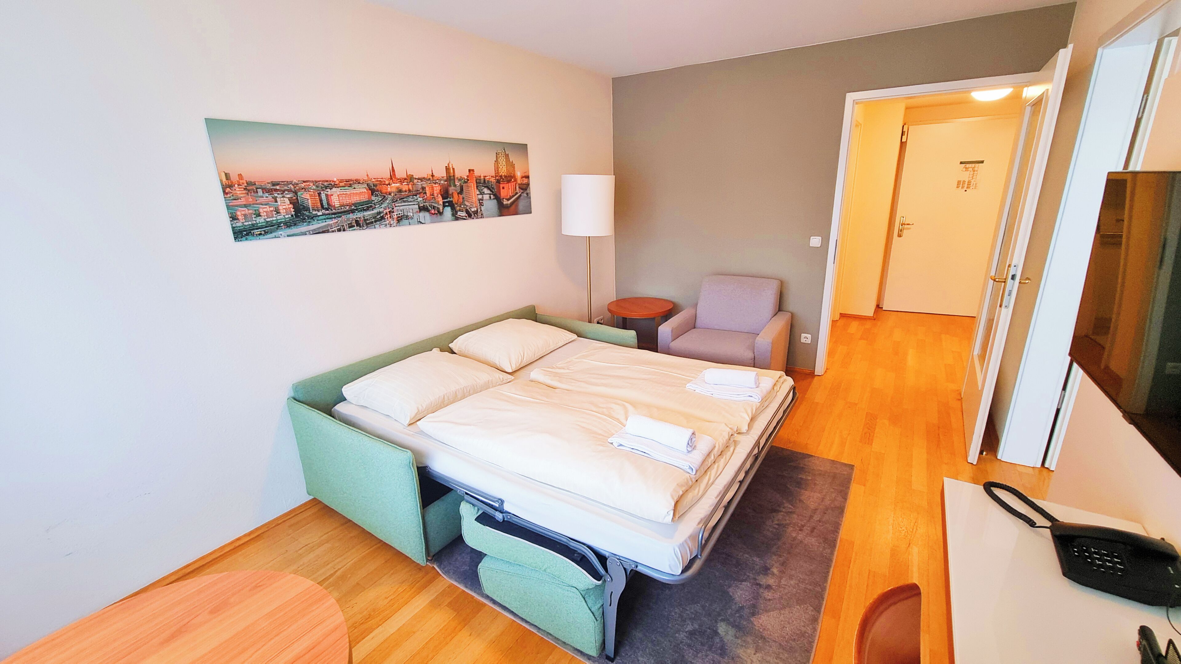 Habitación de hotel con una cama, una silla, una mesita, una lámpara y un cuadro de un paisaje urbano en la pared.
