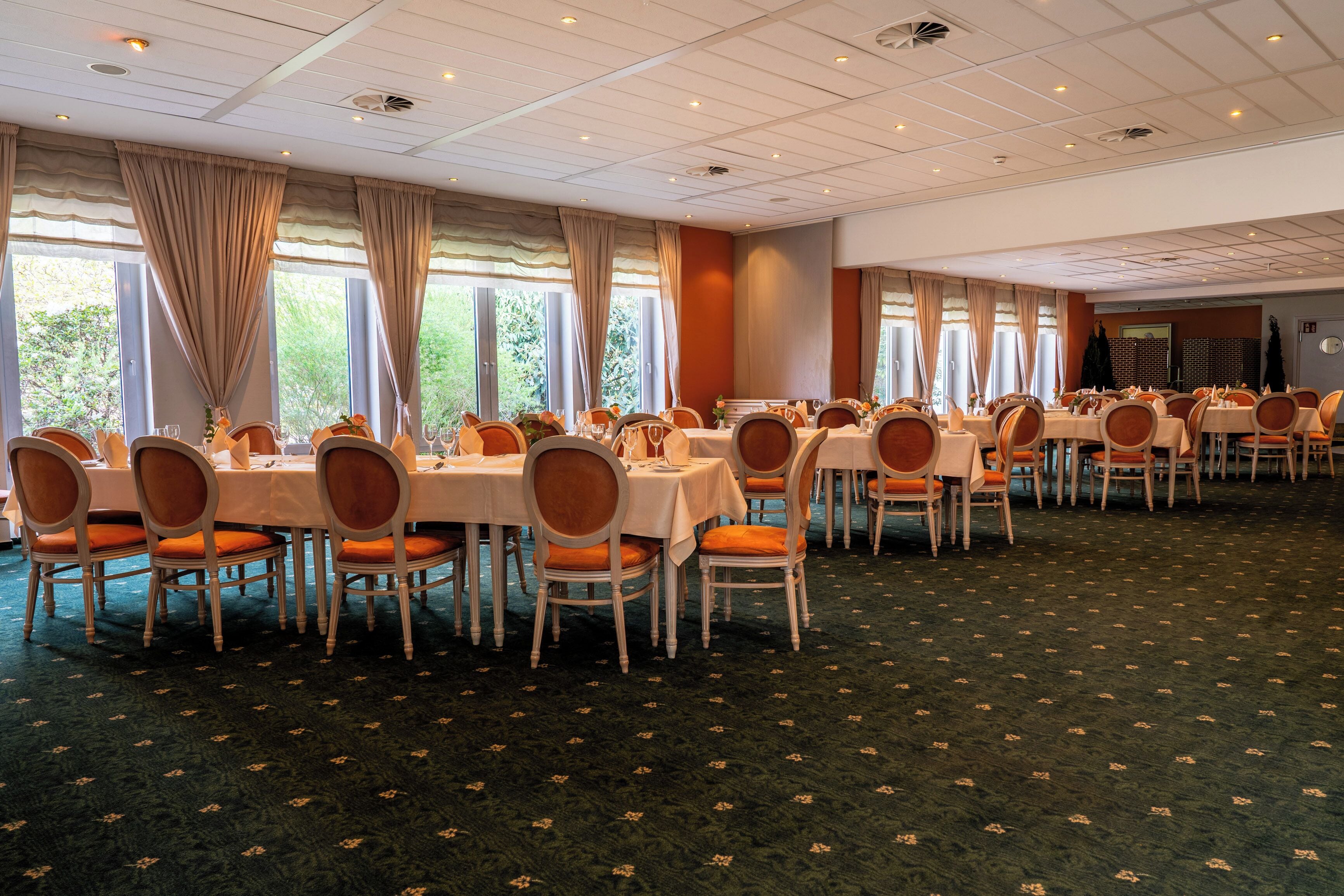 A banquet hall with multiple round tables and chairs, large windows with curtains, and a patterned carpet.