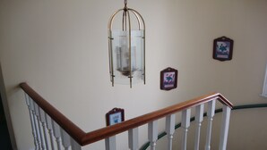 Una escalera con balaustres blancos y barandilla de madera que conduce a una plataforma con un colgador de luces y cuadros enmarcados en la pared.