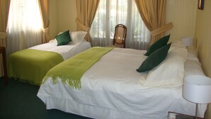 Habitación de hotel con dos camas, cortinas color crema, una silla de madera y una lámpara.