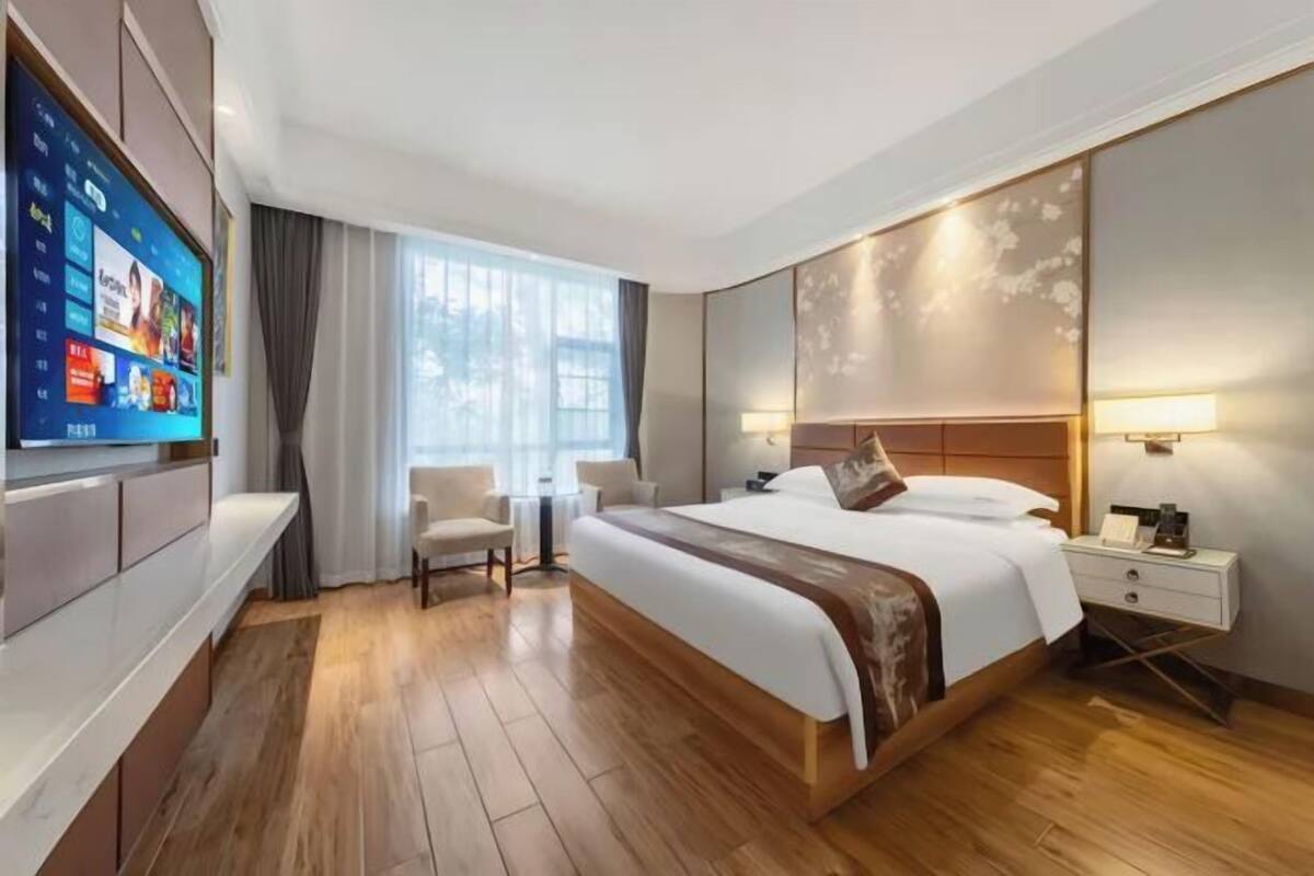 A modern hotel room with a large bed, a TV mounted on the wall, a desk with a lamp, and a view of the outdoors through the window.