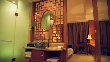 Una habitación de hotel con un separador de madera, un espejo redondo, un lavabo con un cuenco azul, una cama y un escritorio con una lámpara.