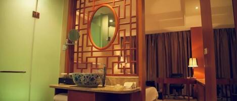A hotel room with a wooden partition, a round mirror, a sink with a blue bowl, a bed, and a desk with a lamp.