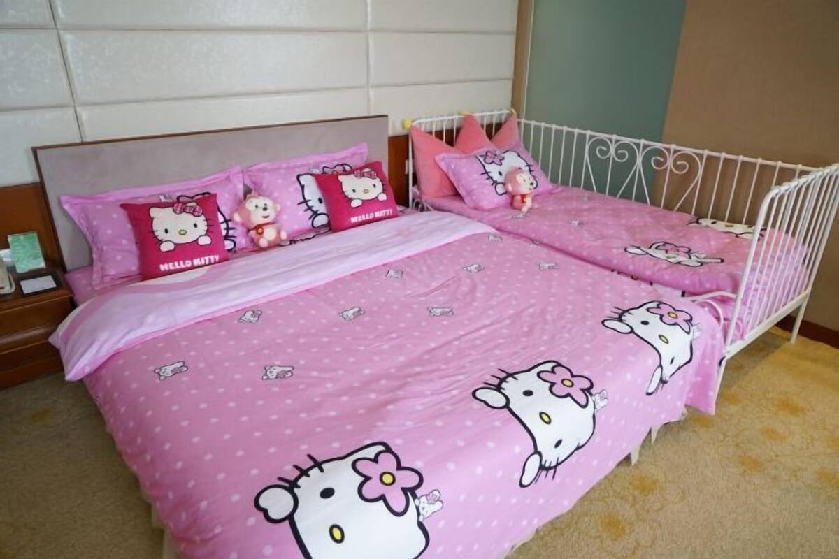 Cama doble con ropa de cama rosa con diseños de Hello Kitty, cabecera metálica blanca y una mesita de noche con un objeto verde.