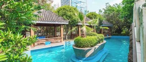 A swimming pool with a small bridge, surrounded by greenery and a building in the background.