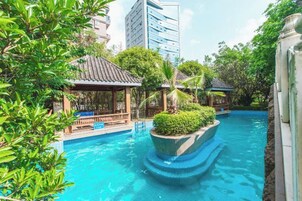 Una piscina con un pequeño puente, rodeada de vegetación y un edificio al fondo.