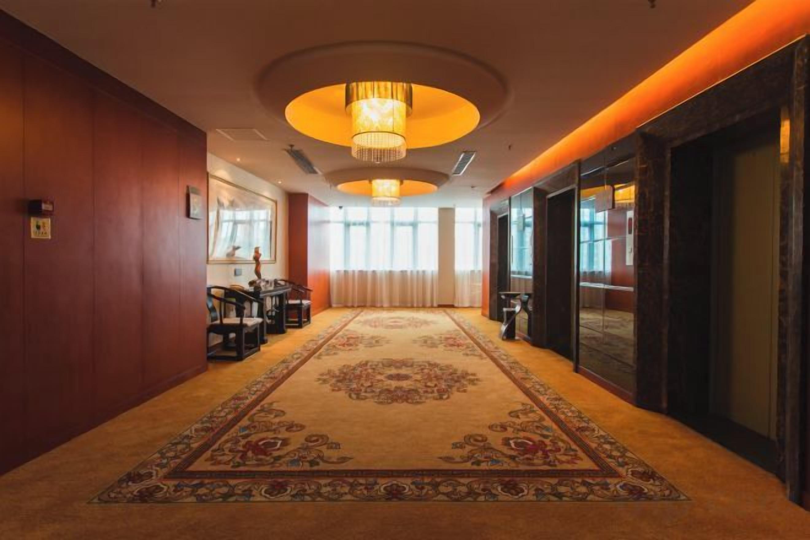 Un couloir d’hôtel avec un sol recouvert de moquette, un lustre et une grande fenêtre.