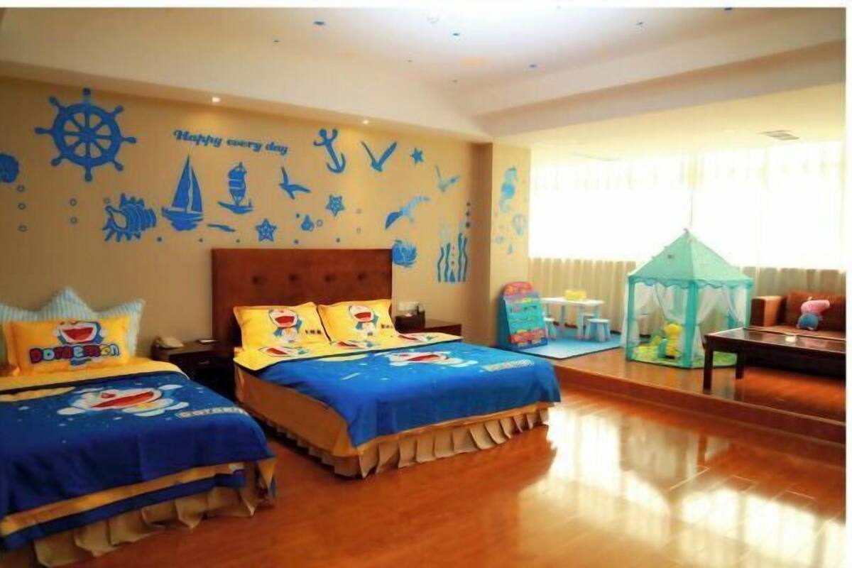Habitación de hotel con dos camas, decoración temática náutica, una ventana con cortinas y una pequeña zona de juegos.