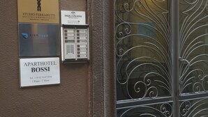 A building entrance with a wrought iron gate, a sign for 'Aparthotel Bossi' with contact details, and multiple business signs including 'Studio Ferraretti' and 'Riva Reno'. - ApartHotel Bossi (Milan)