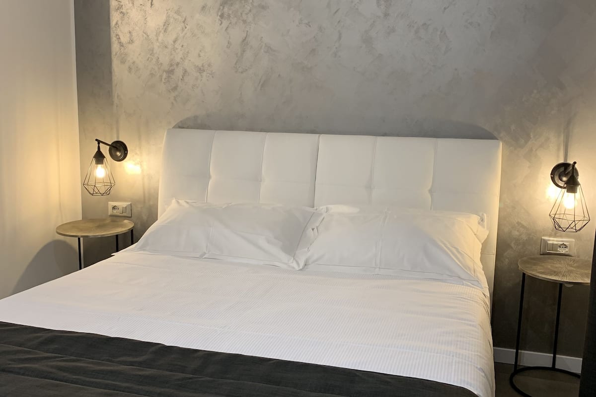Una cama bien tendida con sábanas blancas y un cabecero oscuro, flanqueada por dos mesitas de noche con lámparas de pared.