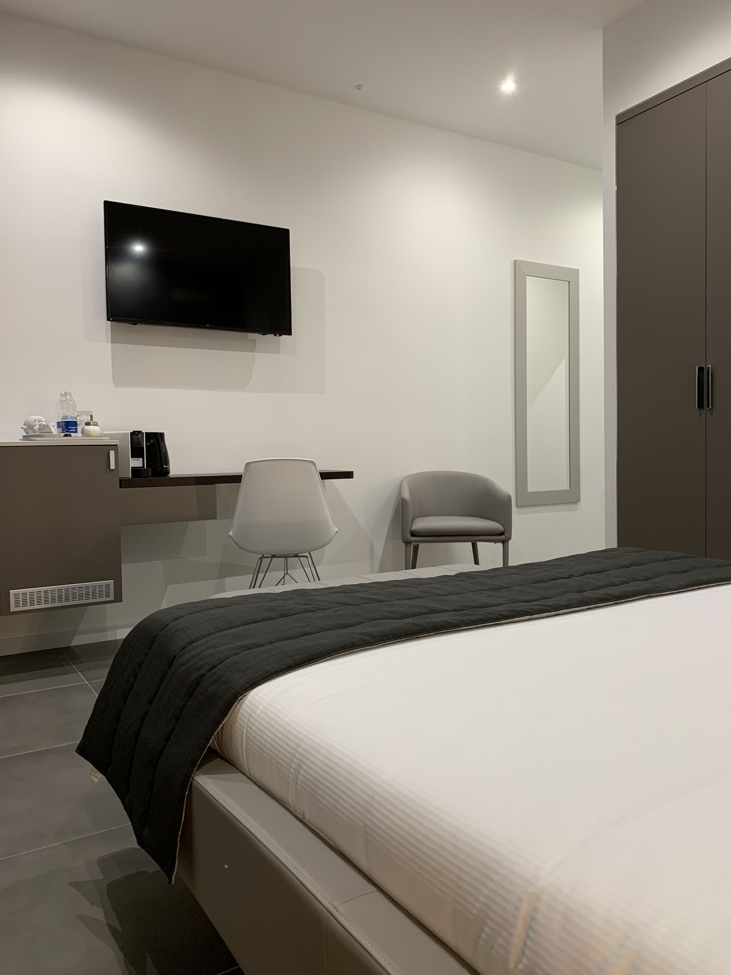 Una habitación de hotel moderna con una cama grande, un televisor de pantalla plana montado en la pared, un escritorio pequeño con cafetera, una silla y un armario.