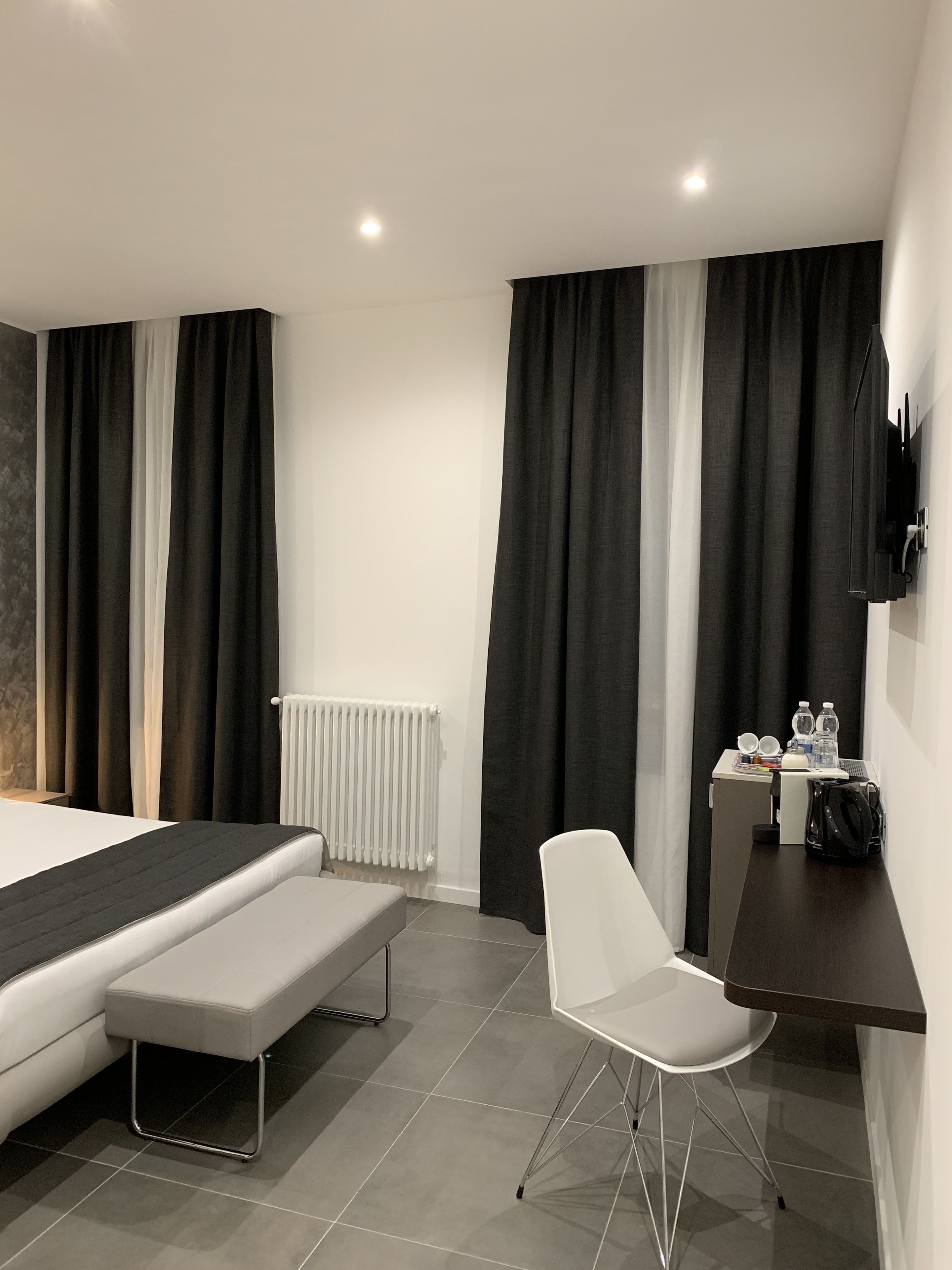 Une chambre d’hôtel moderne équipée d’un lit, d’une banquette, d’une chaise blanche, d’un bureau et d’un téléviseur fixé au mur.
