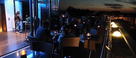 An outdoor dining area with tables and chairs, a glass barrier, and a view of the cityscape.