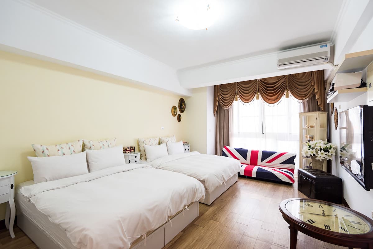 A hotel room with two beds, a large window with curtains, a flat-screen TV, a bedside table, and a wall-mounted air conditioning unit.