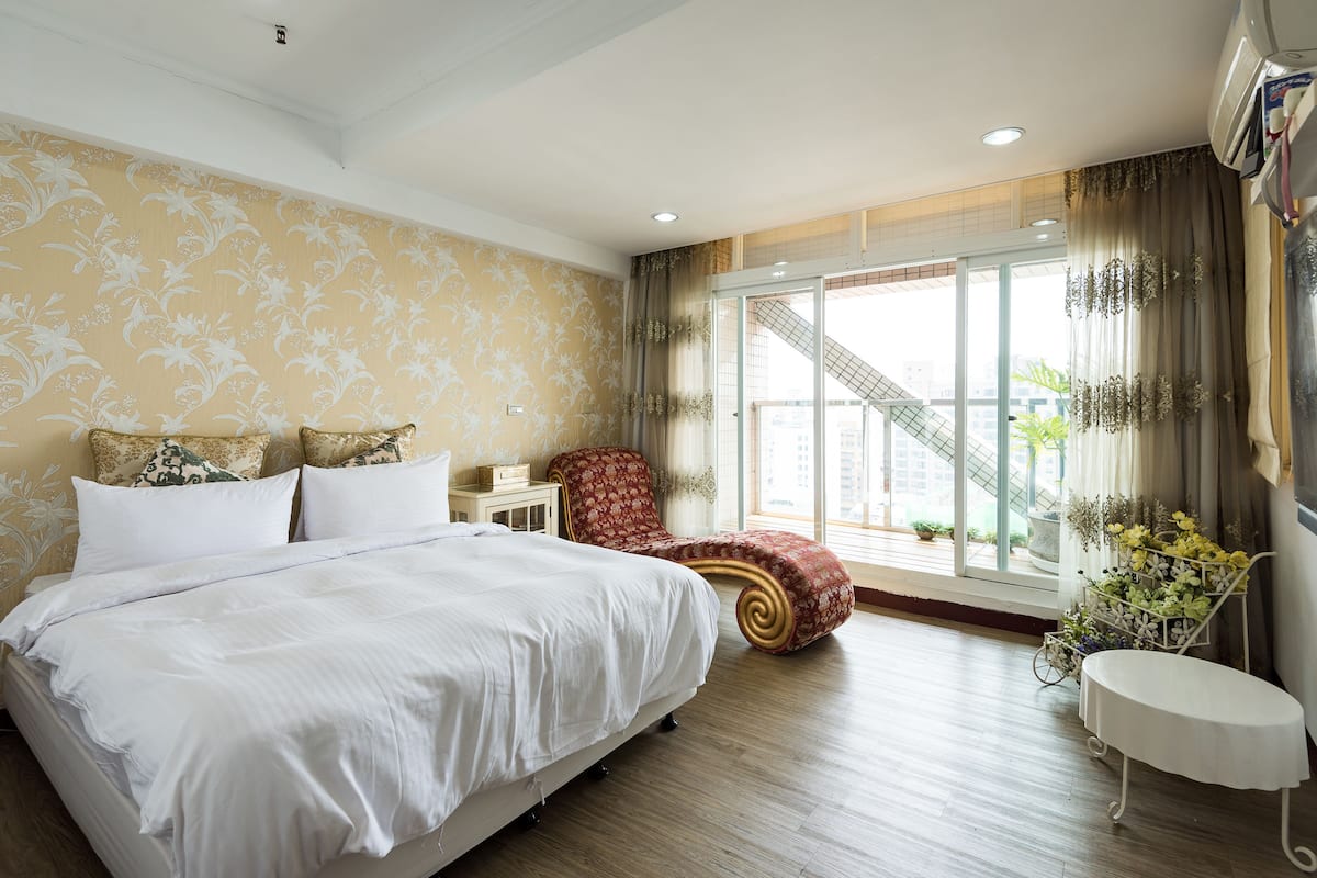 A bedroom with a large bed, a red chair, a small table, and a window with a view of buildings.