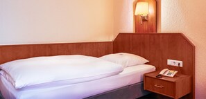 A single bed with a wooden headboard, a bedside table with a telephone, and a wall-mounted light fixture. - Hotel Muenkel (Hannover)