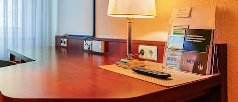 A hotel room with a wooden desk, a lamp, a remote control, and a guidebook on the table.