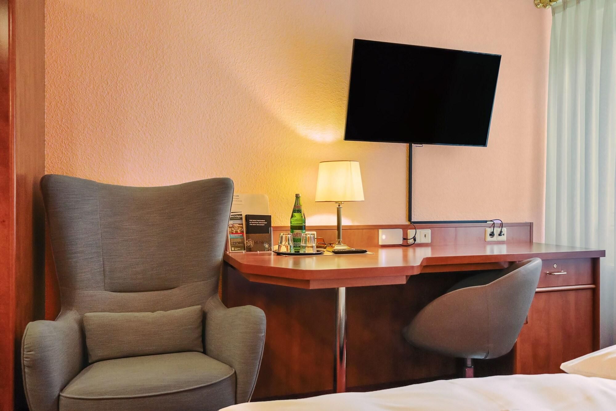 A hotel room with a large armchair, a desk with a lamp, a television mounted on the wall, and a bed.