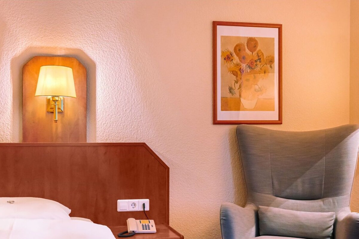 A hotel room with a bed, a bedside lamp, a telephone, a chair, and a painting on the wall.