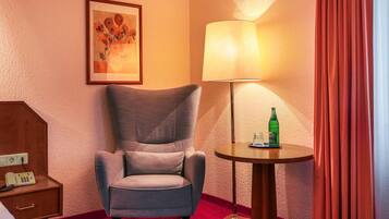 A hotel room with a grey armchair, a bedside table with a telephone, a lamp, a bottle of water, and a framed artwork on the wall.