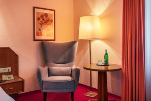 A hotel room with a grey armchair, a bedside table with a telephone, a lamp, a bottle of water, and a framed artwork on the wall. - Hotel Muenkel (Hannover)