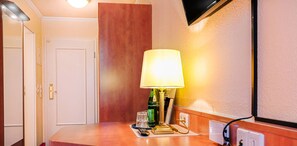 A hotel room with a wooden desk, a lamp, a remote control, a book, and a glass. - Hotel Muenkel (Hannover)