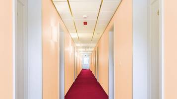 A long, narrow hallway with cream-colored walls and a red carpet, leading to a door at the end.