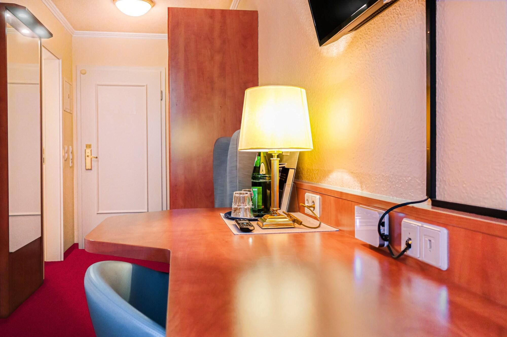 Una habitación de hotel con un escritorio de madera, una lámpara, una botella, un vaso y un control remoto.