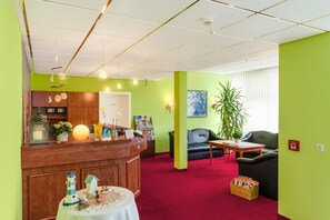 A reception area with a wooden counter, a small table with a decorative item, a reception desk, a black sofa, a red carpet, and a large plant. - Hotel Muenkel (Hannover)