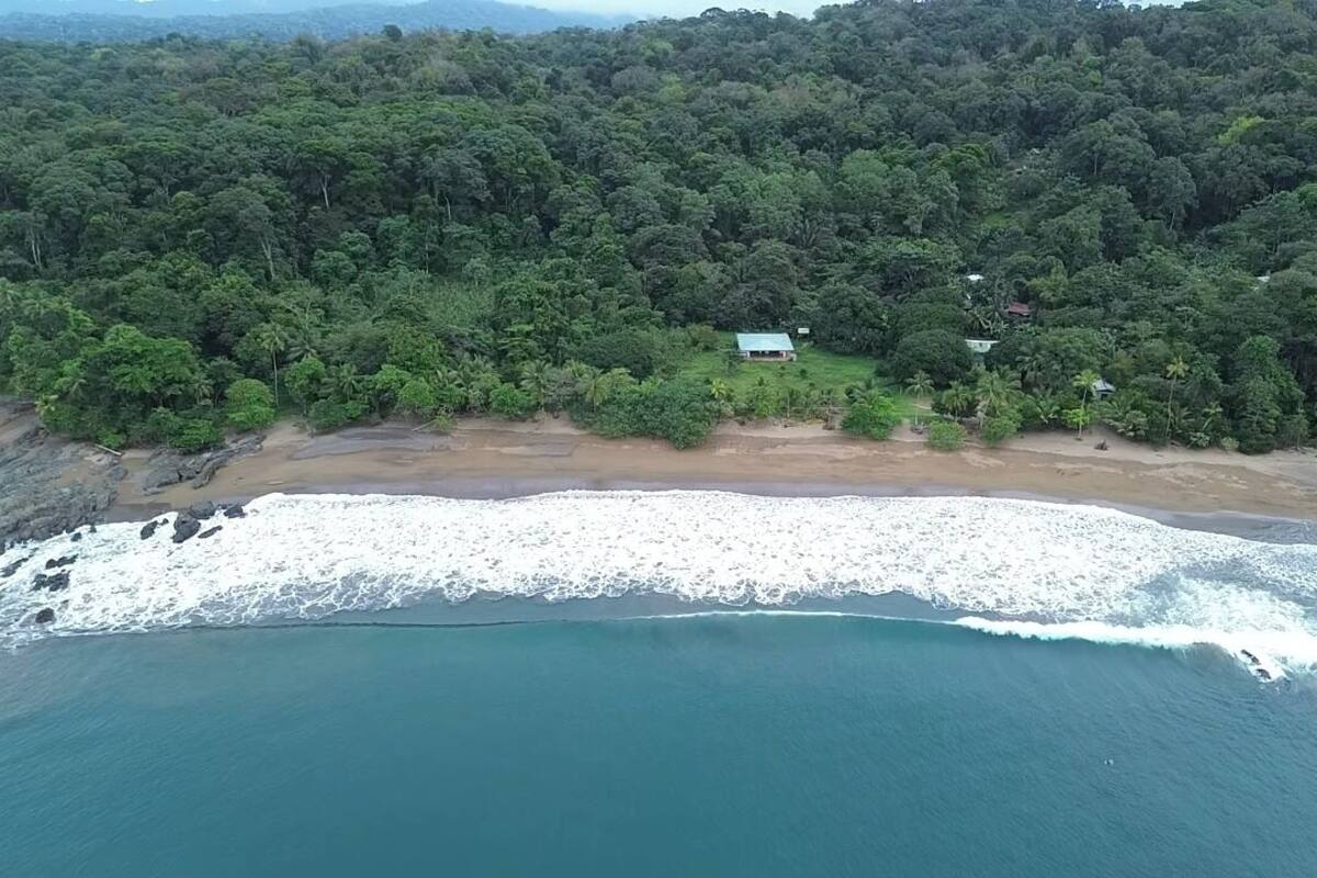An aerial view of a coastal area with a beach, forested hills, and a small building with a green roof.