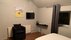 A bedroom with a bed, a black armchair, a painting, a TV, a shelf with cups and a kettle, and a window with curtains. - Halfway House (Blackwood)