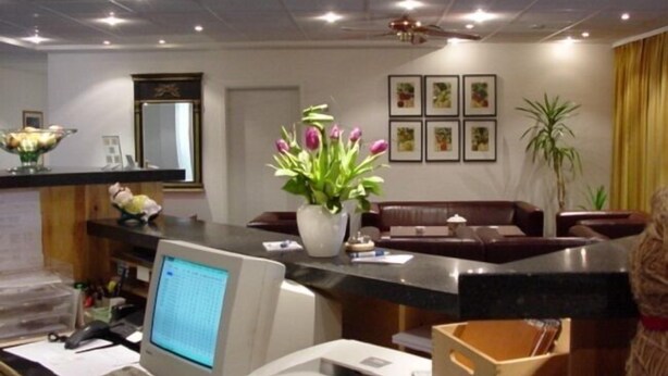 A hotel reception area with a computer, a desk, a vase of flowers, framed pictures on the wall, and a plant. - Neustädter Hof Hotel Garni (Neustadt am Ruebenberge)