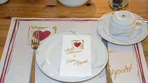A table set with a white plate, a gold fork, a gold spoon, a white napkin with a heart design, and a white teacup and saucer. - Neustädter Hof Hotel Garni (Neustadt am Ruebenberge)