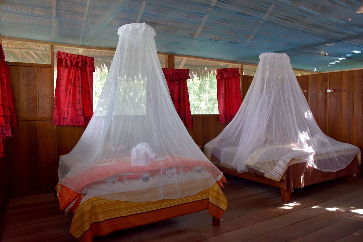 Dos camas de madera con mosquiteros blancos, cortinas rojas y techo de madera.