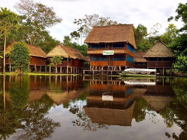 Ein traditionelles Holzhaus mit einem Strohdach, umgeben von ruhigem Wasser, und ein kleines Boot liegt in der Nähe.