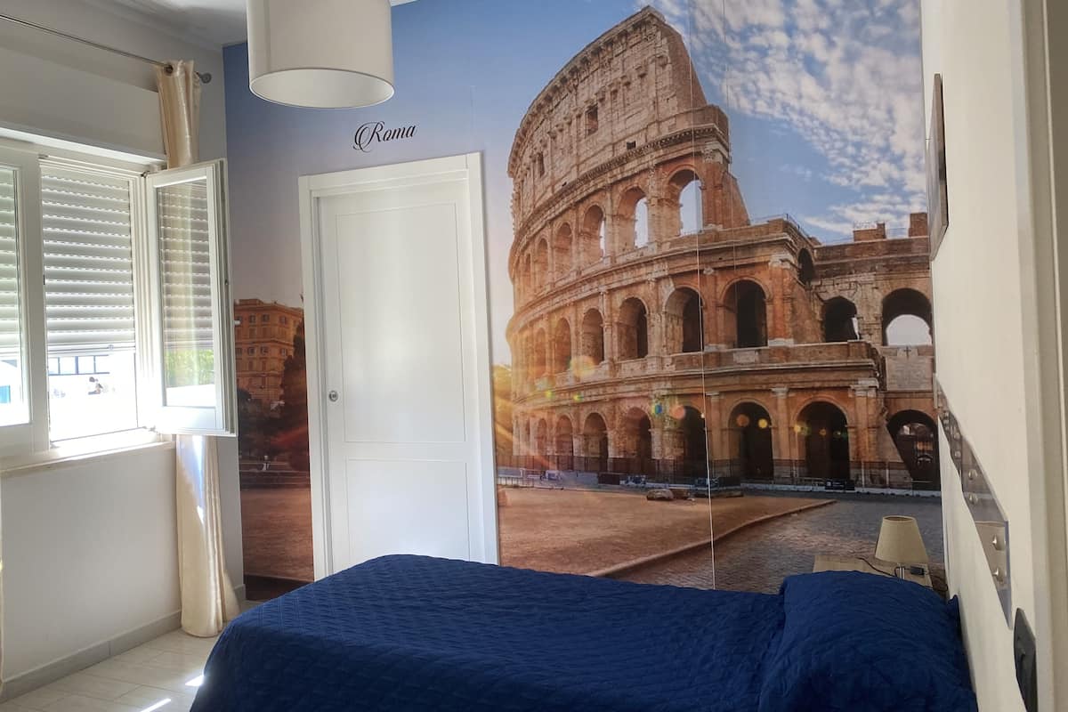 A bedroom with a large mural of the Colosseum, a white door, and a window with blinds.