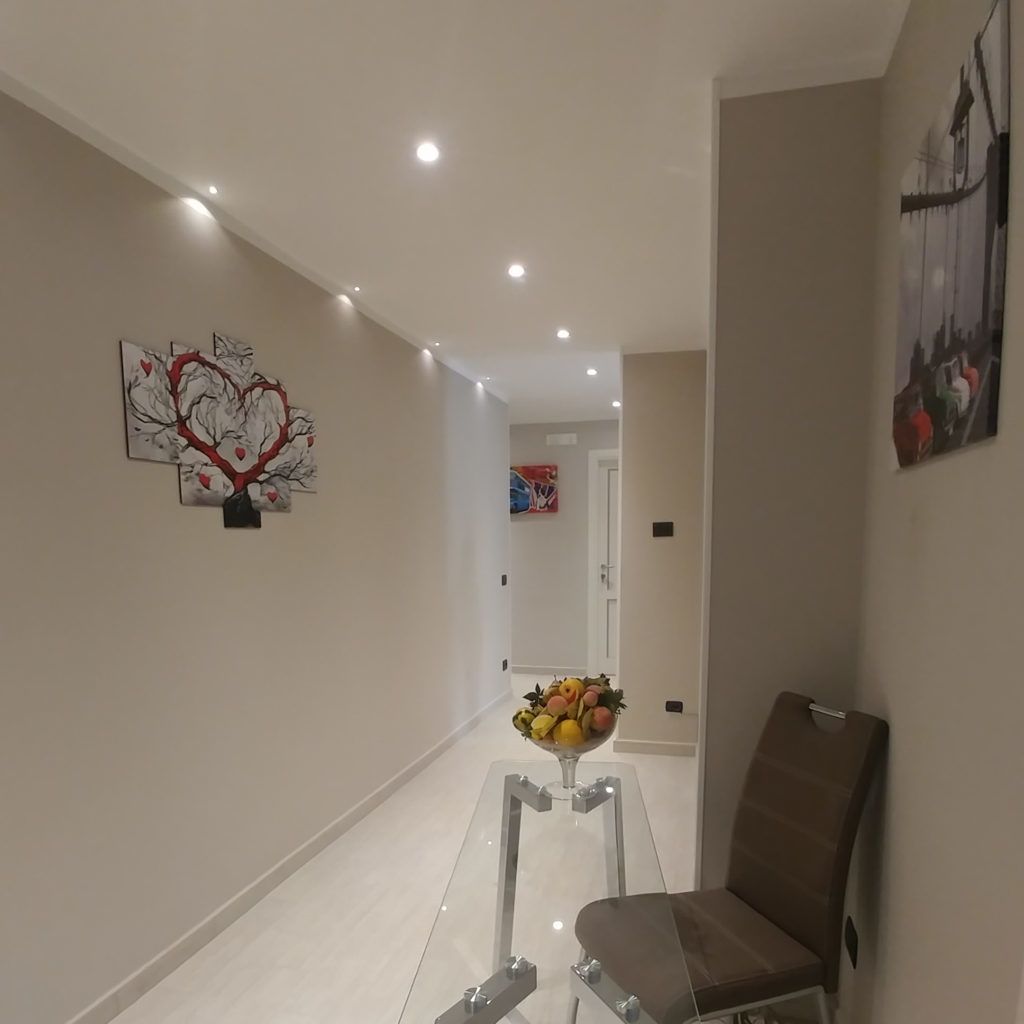 A hallway with a modern design, featuring a glass table with a fruit bowl, a brown chair, and artwork on the walls.