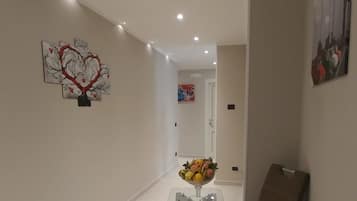 A hallway with a modern design, featuring a glass table with a fruit bowl, a brown chair, and artwork on the walls.
