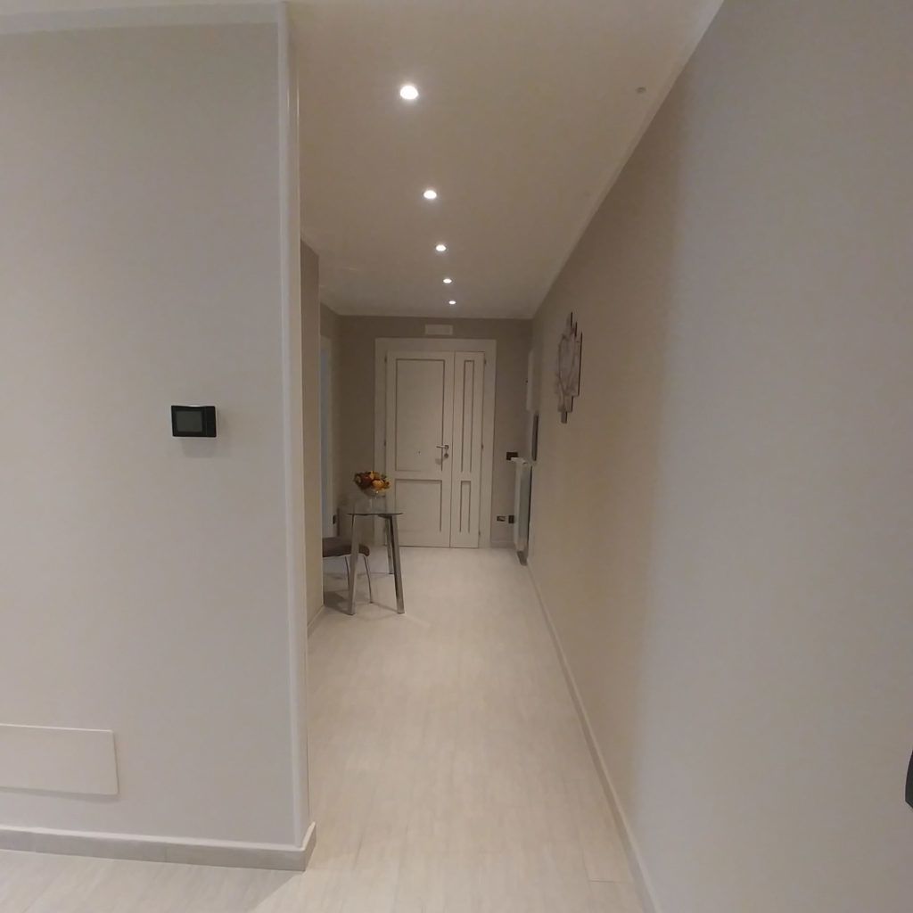 A hallway with a door at the end, a small table with a vase of flowers, and recessed lighting on the ceiling.