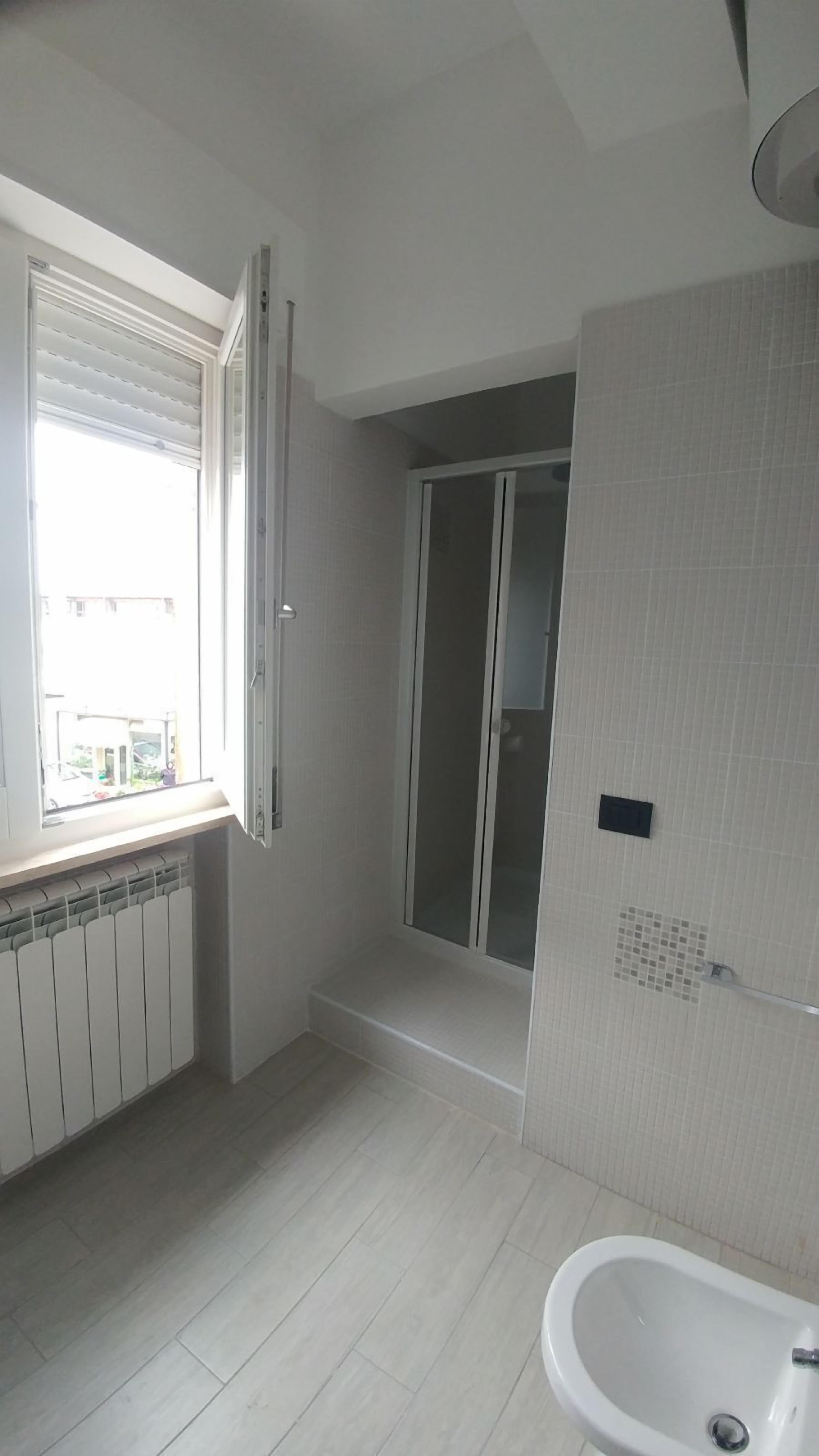 A bathroom with a toilet, a sink, a window with blinds, a shower area, and a tiled wall.