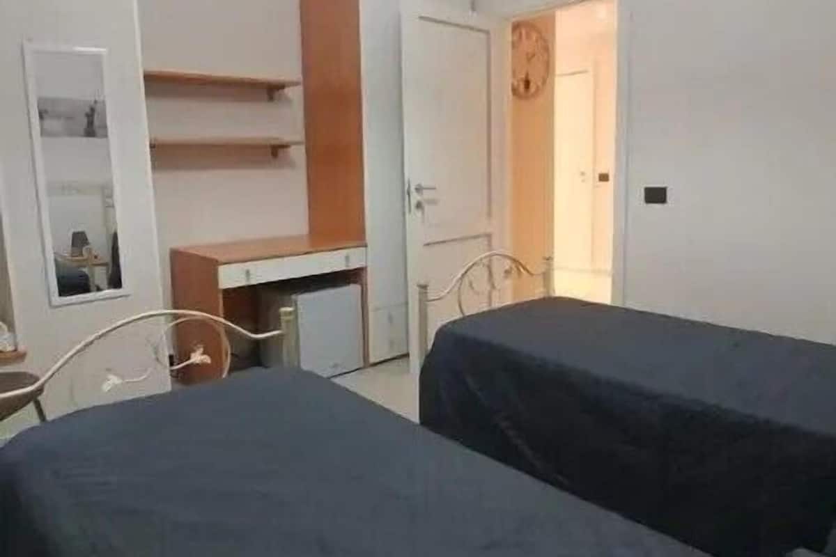 A small, well-lit room with two beds, a desk, and a wardrobe.